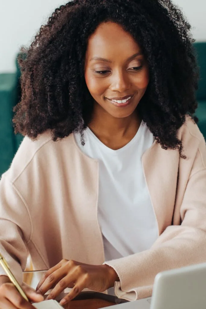 A woman smiling while writing and using a laptop, representing personal coaching for life management